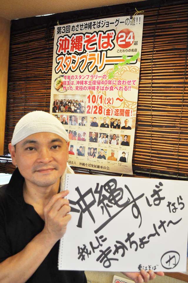 「沖縄そばスタンプラリー」のポスターの前でポーズを取る屋比久さん
